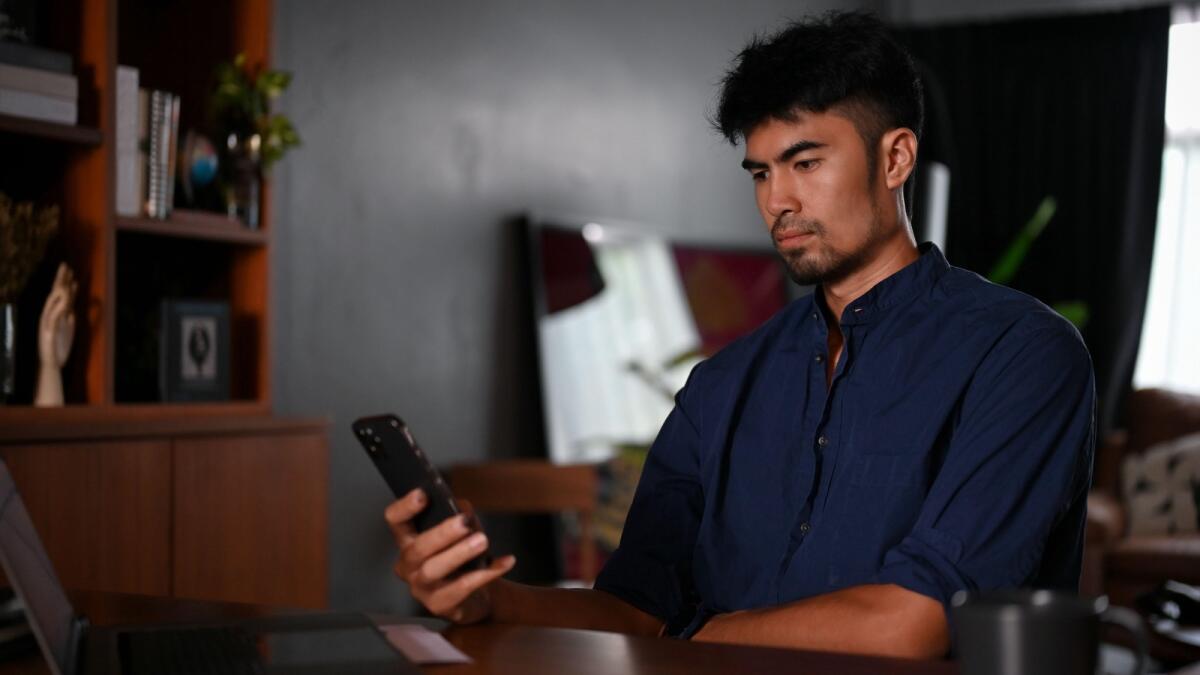 Handsome millennial Asian man in his living room, using a smartphone, scrolling on social media platform, chatting with someone.